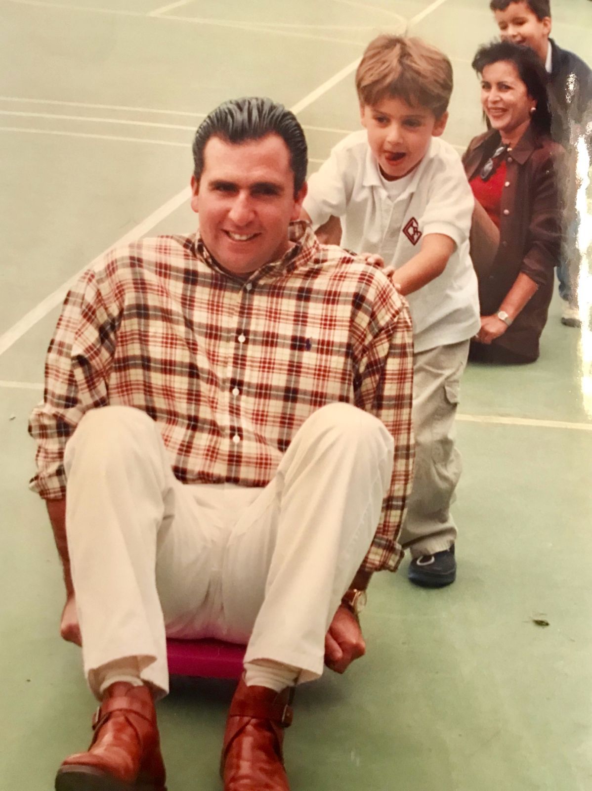 A family photograph from the 1990s showing Roberto Arzú being pushed on a scooter by a child at a school event